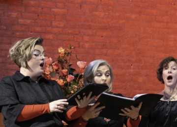 Een koor van acht volwassen zangers, gekleed in zwart met oranje accenten, zingt tegen een bakstenen muur.