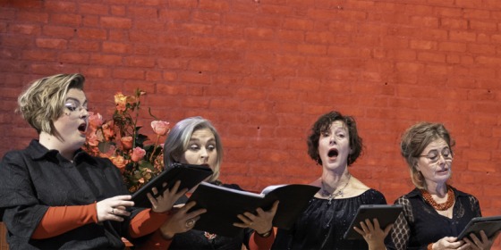 Een koor van acht volwassen zangers, gekleed in zwart met oranje accenten, zingt tegen een bakstenen muur.