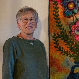 Oudere vrouw met bril poseert naast een kleurrijk wandtapijt met bloemen in een vaas op de muur.