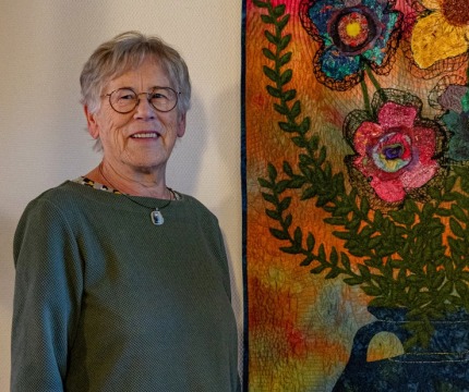 Oudere vrouw met bril poseert naast een kleurrijk wandtapijt met bloemen in een vaas op de muur.