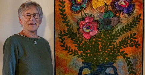 Oudere vrouw met bril poseert naast een kleurrijk wandtapijt met bloemen in een vaas op de muur.