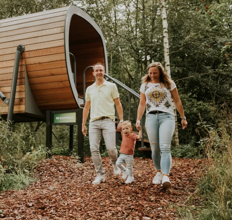 Gezin wandelt samen in het bos bij een modern houten boomhutverblijf op een verhoogde constructie.