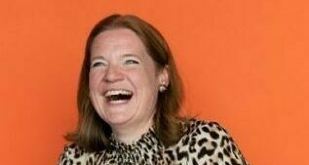 Vrouw met rood haar in panterprint blouse lacht breed en zit aan tafel met een groot groen boek voor een oranje muur.