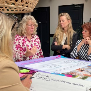 Vijf vrouwen zitten rond een tafel en bespreken notities tijdens een groepssessie in een moderne ruimte.