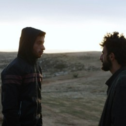 Twee mannen staan tegenover elkaar in een open landschap bij zonsopgang of zonsondergang, in gesprek.