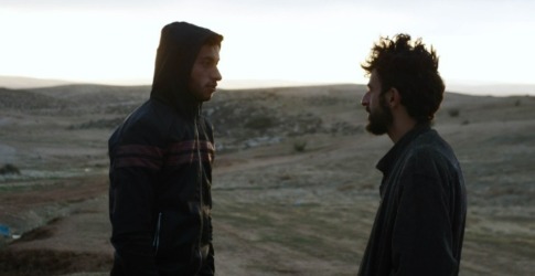 Twee mannen staan tegenover elkaar in een open landschap bij zonsopgang of zonsondergang, in gesprek.