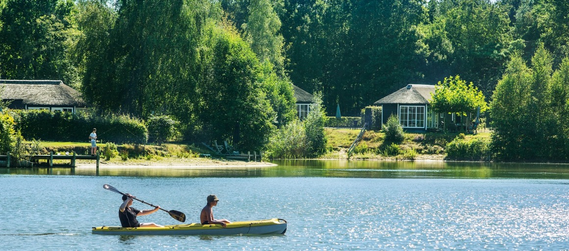 Twee mensen kanoën op een rustig meer met vakantiehuisjes en groene bomen op de achtergrond in de zomer.