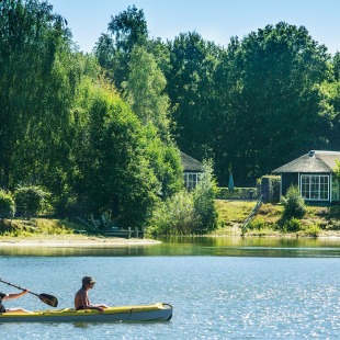 Twee mensen kanoën op een rustig meer met vakantiehuisjes en groene bomen op de achtergrond in de zomer.
