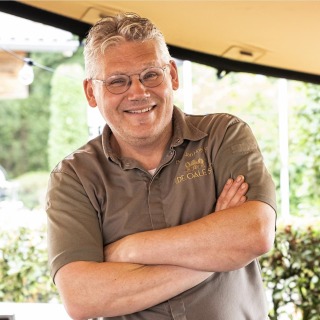 Man met bril en grijs haar, gekleed in een bruin overhemd met opdruk, lacht en staat met gekruiste armen.