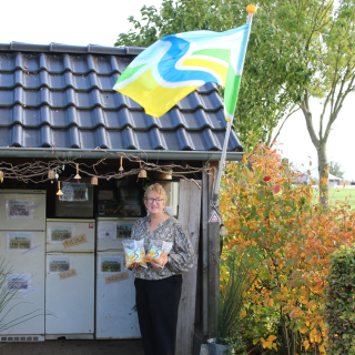 Vrouw staat voor een boerderijwinkel met zakken producten, kleurrijke vlag wappert en bomen op de achtergrond.
