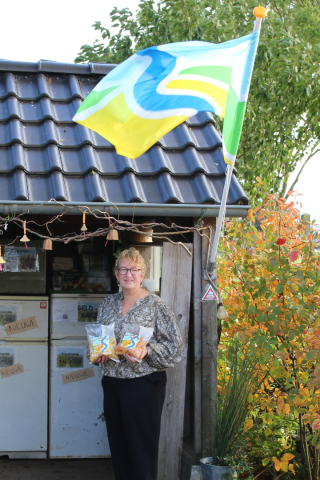 Vrouw staat voor een boerderijwinkel met zakken producten, kleurrijke vlag wappert en bomen op de achtergrond.