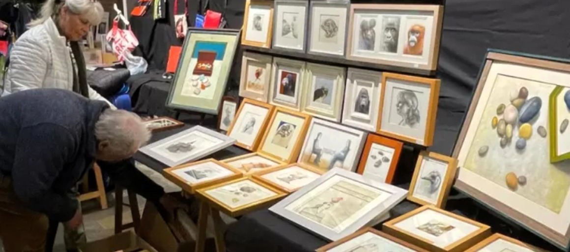 Twee mensen bekijken ingelijste kunstwerken op een kunstmarkt, met tekeningen en schilderijen aan tafels.