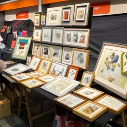 Twee mensen bekijken ingelijste kunstwerken op een kunstmarkt, met tekeningen en schilderijen aan tafels.