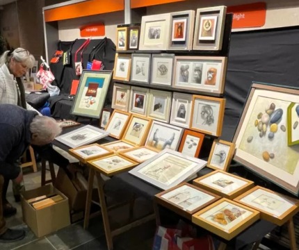 Twee mensen bekijken ingelijste kunstwerken op een kunstmarkt, met tekeningen en schilderijen aan tafels.