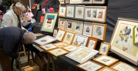 Twee mensen bekijken ingelijste kunstwerken op een kunstmarkt, met tekeningen en schilderijen aan tafels.