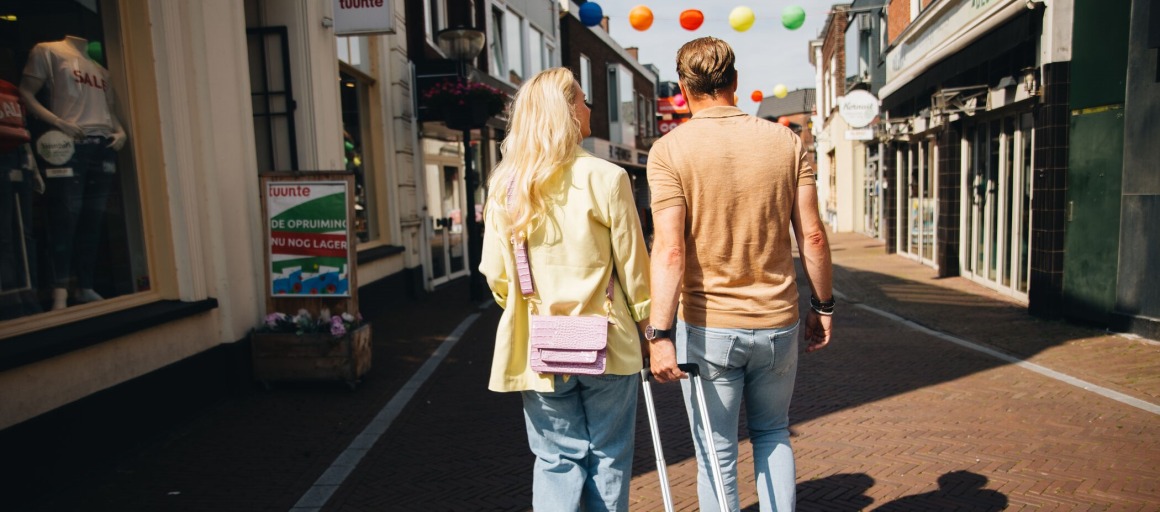 Twee mensen lopen hand in hand door een winkelstraat met een turquoise koffer op een zonnige dag.