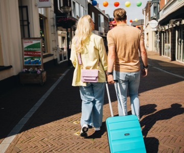 Twee mensen lopen hand in hand door een winkelstraat met een turquoise koffer op een zonnige dag.