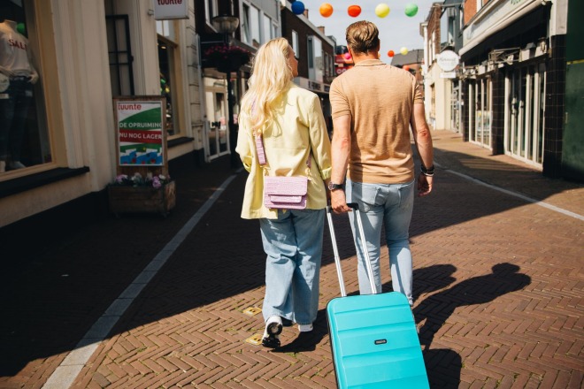Twee mensen lopen hand in hand door een winkelstraat met een turquoise koffer op een zonnige dag.