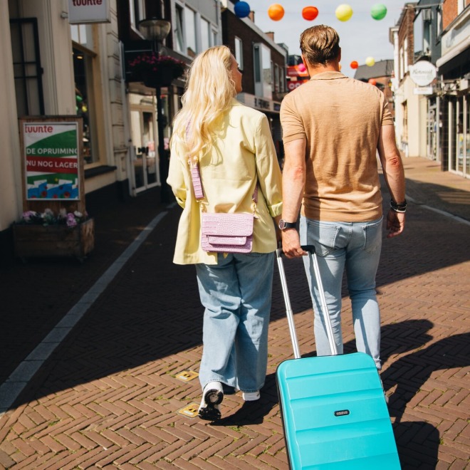 Twee mensen lopen hand in hand door een winkelstraat met een turquoise koffer op een zonnige dag.