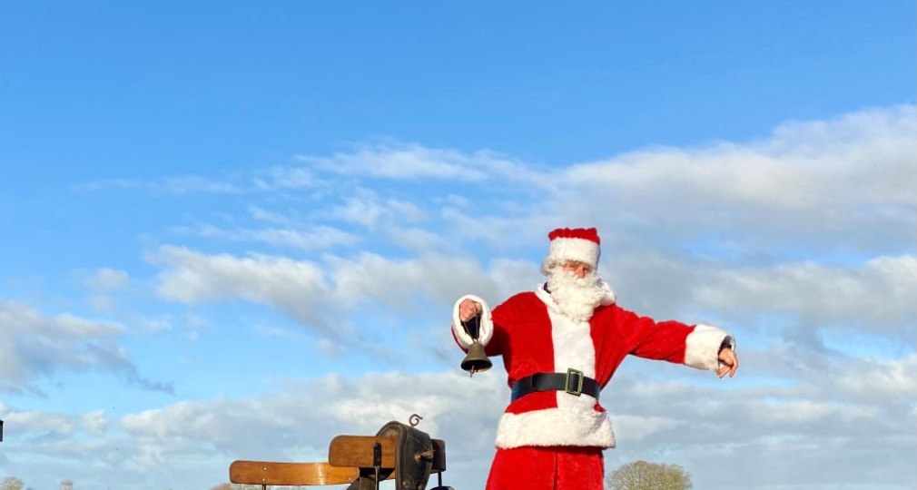 Een persoon verkleed als kerstman staat op een houten wagen in een open veld onder een blauwe lucht.