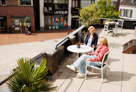 Twee vrouwen zitten op een zonnig buitenterras met uitzicht op een winkelstraat in een gezellig centrum.