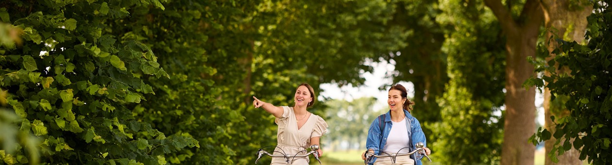 Twee vrouwen fietsen samen op een bosrijk pad, lachend en genietend van de natuur om hen heen.