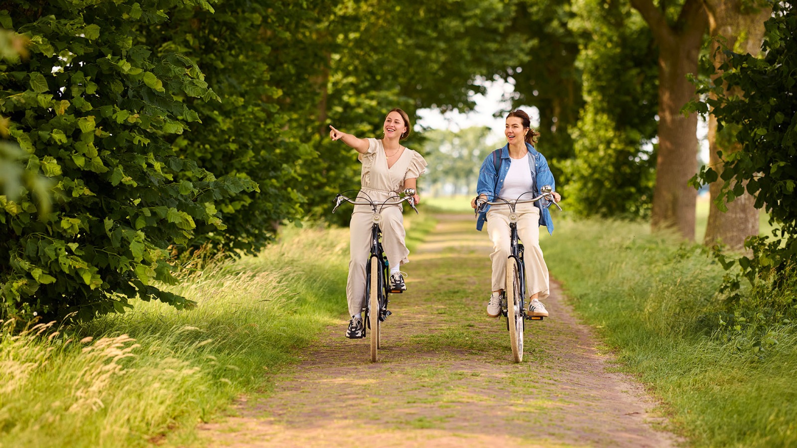Twee vrouwen fietsen samen op een bosrijk pad, lachend en genietend van de natuur om hen heen.