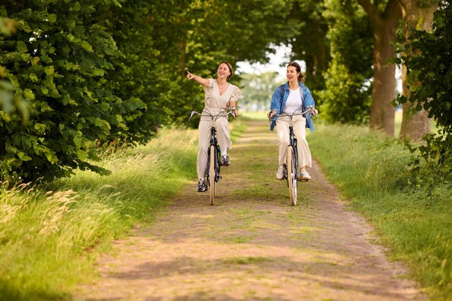 Twee vrouwen fietsen samen op een bosrijk pad, lachend en genietend van de natuur om hen heen.