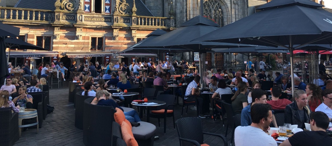 Drukke terrassen vol mensen bij een historisch gebouw in de avondzon op een stadsplein in Nederland.