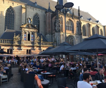 Drukke terrassen vol mensen bij een historisch gebouw in de avondzon op een stadsplein in Nederland.