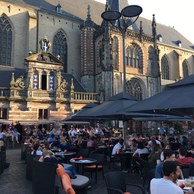 Drukke terrassen vol mensen bij een historisch gebouw in de avondzon op een stadsplein in Nederland.