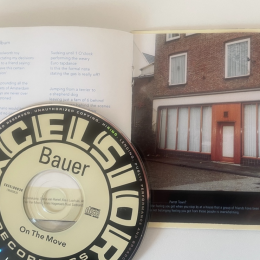 CD van Bauer met het album 'On The Move', boekje open bij een foto van een hoekpand met gesloten gordijnen.