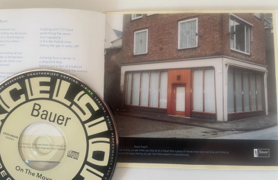 CD van Bauer met het album 'On The Move', boekje open bij een foto van een hoekpand met gesloten gordijnen.