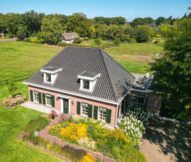 Luchtfoto van een traditioneel Nederlands huis met zwart dak, omgeven door groene weiden en tuinen.