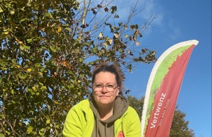 Persoon met bril in gele jas zit buiten op krukje met een drankje voor een Vertwenz vlag, omgeven door groen.