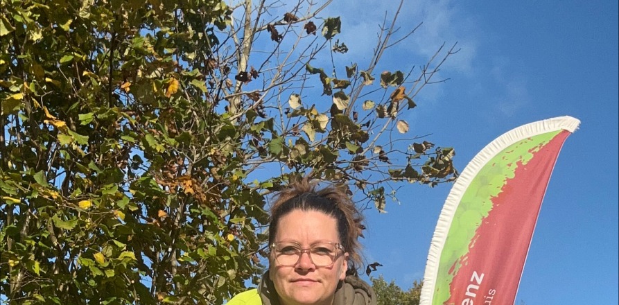 Persoon met bril in gele jas zit buiten op krukje met een drankje voor een Vertwenz vlag, omgeven door groen.