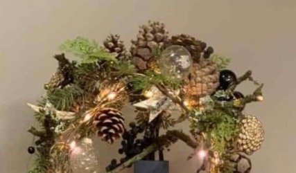 Weihnachtliche Dekoration mit Tannenzweigen, Zapfen, Kugeln und Lichtern auf einem schwarzen Sockel.