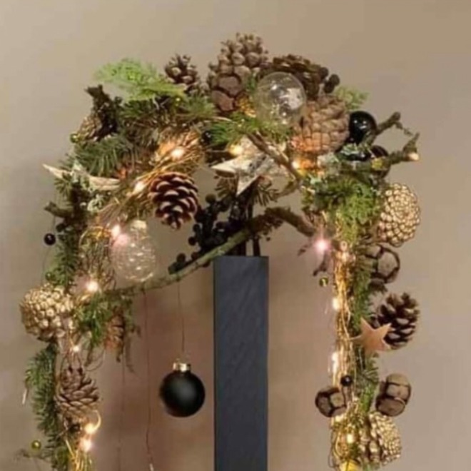 Weihnachtliche Dekoration mit Tannenzweigen, Zapfen, Kugeln und Lichtern auf einem schwarzen Sockel.