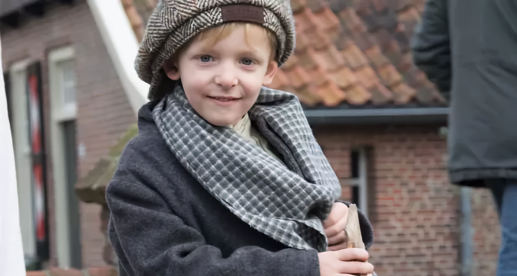 Jonge jongen verkleed in vintage kleren met een pet, sjaal en jas, zittend op een houten stokpaard.