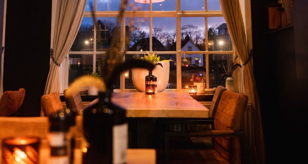 Gezellige restaurantsetting met grote ramen, sfeerverlichting en gedekte tafels tijdens de schemering.