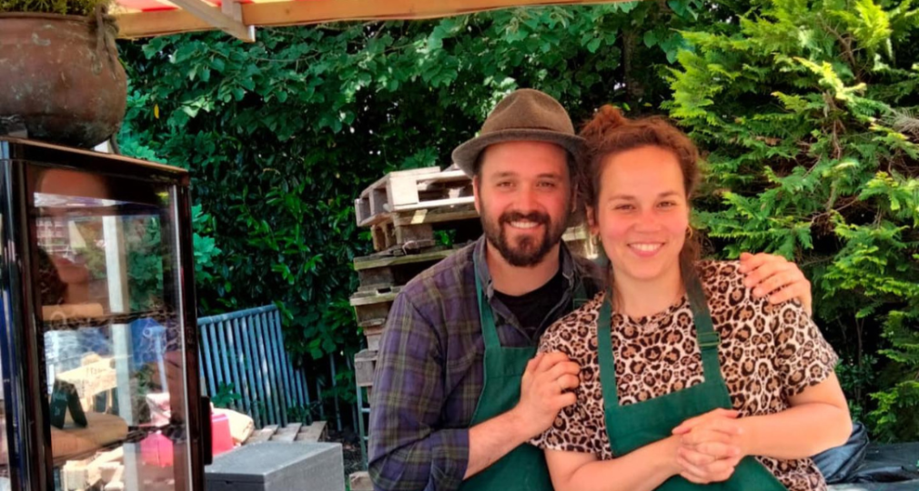 Twee mensen in groene schorten lachen bij een kraam met gebak, taart en desserts op een markt.