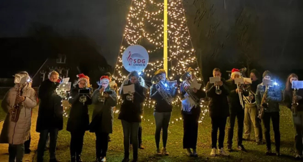 Muzikanten spelen onder een verlichte kerstboom, buiten ’s avonds, met SDG Ommen bord zichtbaar.
