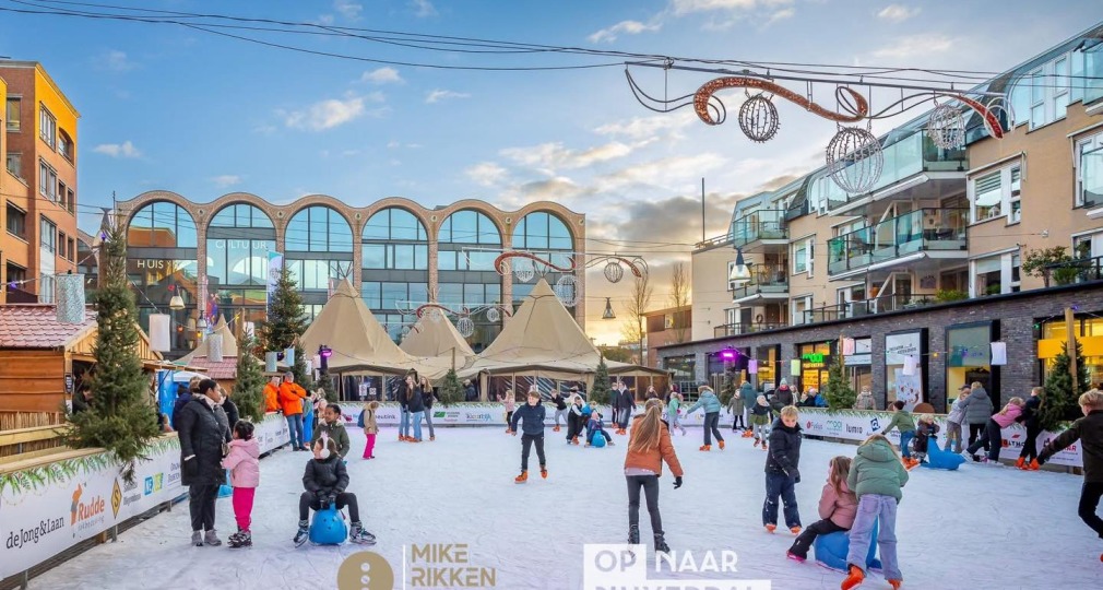 Kinderen en volwassenen schaatsen op een buitenijsbaan in een stadsplein met gezellige winterdecoratie.