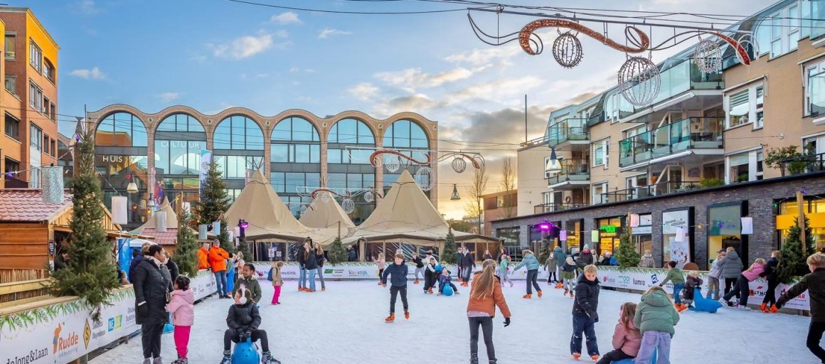 Kinderen en volwassenen schaatsen op een buitenijsbaan in een stadsplein met gezellige winterdecoratie.