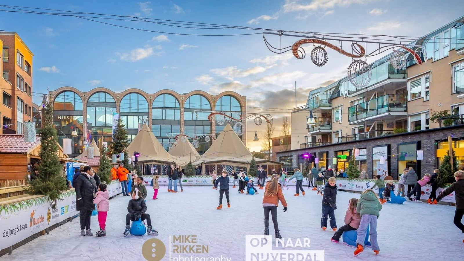 Kinderen en volwassenen schaatsen op een buitenijsbaan in een stadsplein met gezellige winterdecoratie.