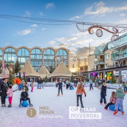 Kinderen en volwassenen schaatsen op een buitenijsbaan in een stadsplein met gezellige winterdecoratie.