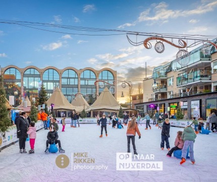 Kinderen en volwassenen schaatsen op een buitenijsbaan in een stadsplein met gezellige winterdecoratie.