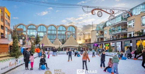 Kinderen en volwassenen schaatsen op een buitenijsbaan in een stadsplein met gezellige winterdecoratie.