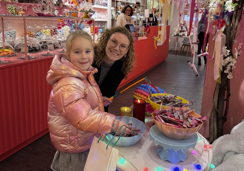 Een jong meisje en een vrouw glimlachen bij een tafel met kleurrijk snoep in een vrolijke winkel tijdens een lampionnentocht.