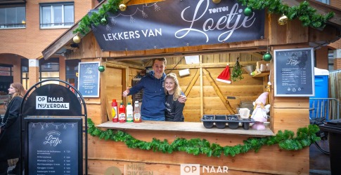 Twee mensen poseren in een houten kerstkraam versierd met groen, gouden ballen en een kerstster.
